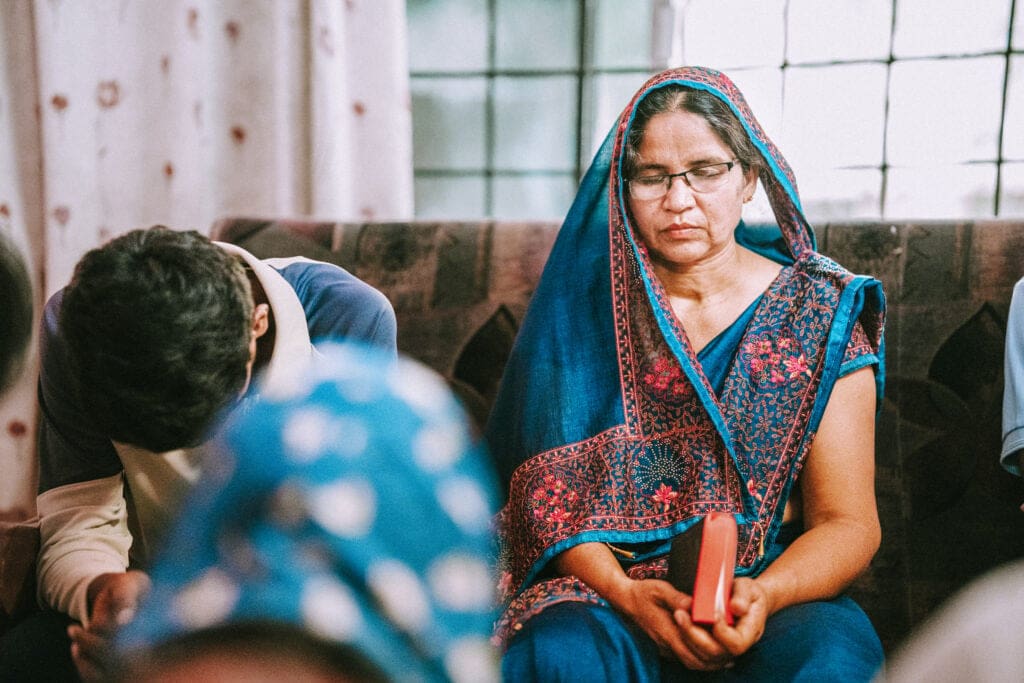 A South Asian woman in a blue sari prays in a small house gathering, eyes closed and holding a small Scripture book.
